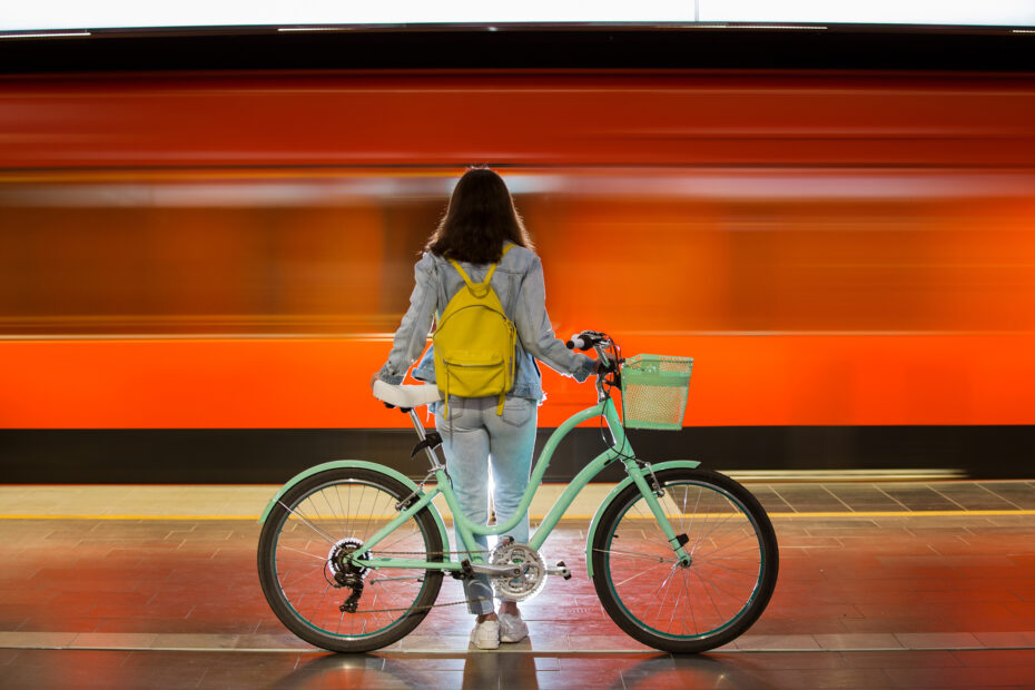 Teenager girl in jeans with yellow backpack and bike standing on metro station, waiting for train, smiling, laughing. Orange train passing by behind the girl. Futuristic subway station. Finland, Espoo